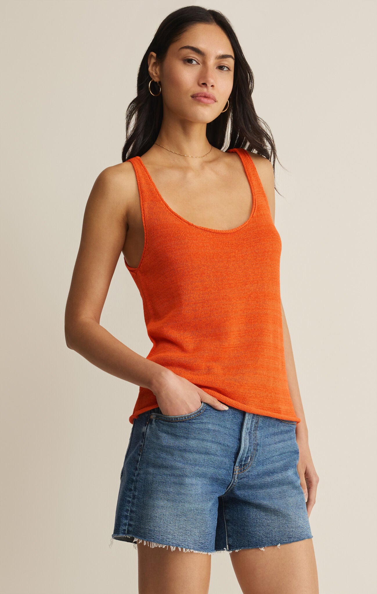 Woman wearing an orange tank top and denim shorts on a beige background