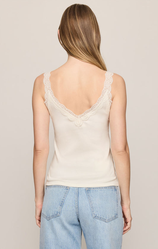 A woman viewed from the back wearing a light cream camisole tank top with lace trim and light blue jeans, set against a plain light beige studio background.