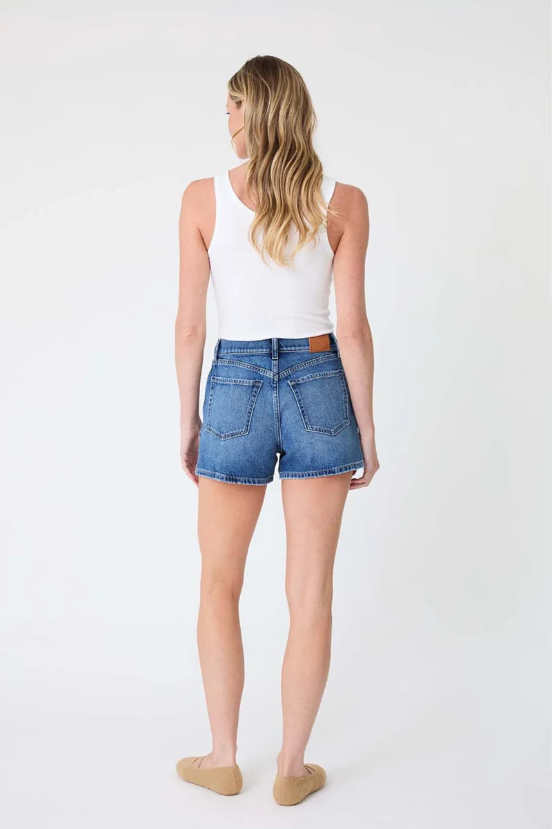 A woman is shown from behind wearing blue denim shorts, a white tank top, and beige flat shoes against a plain white studio background.