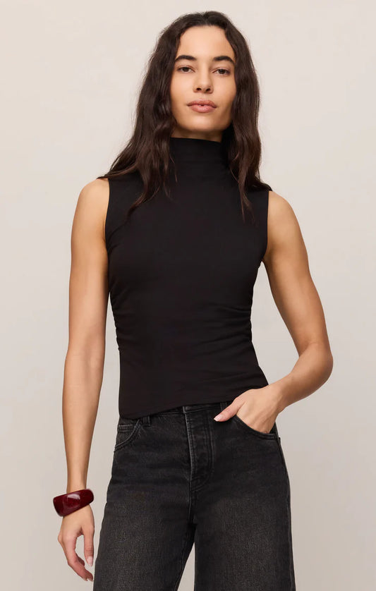 A woman modeling a sleeveless black mock neck top paired with dark gray jeans, standing against a plain light beige studio background.