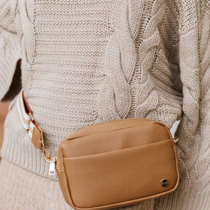 A person wearing a beige knitted sweater with a light camel-colored crossbody sling bag featuring gold hardware.