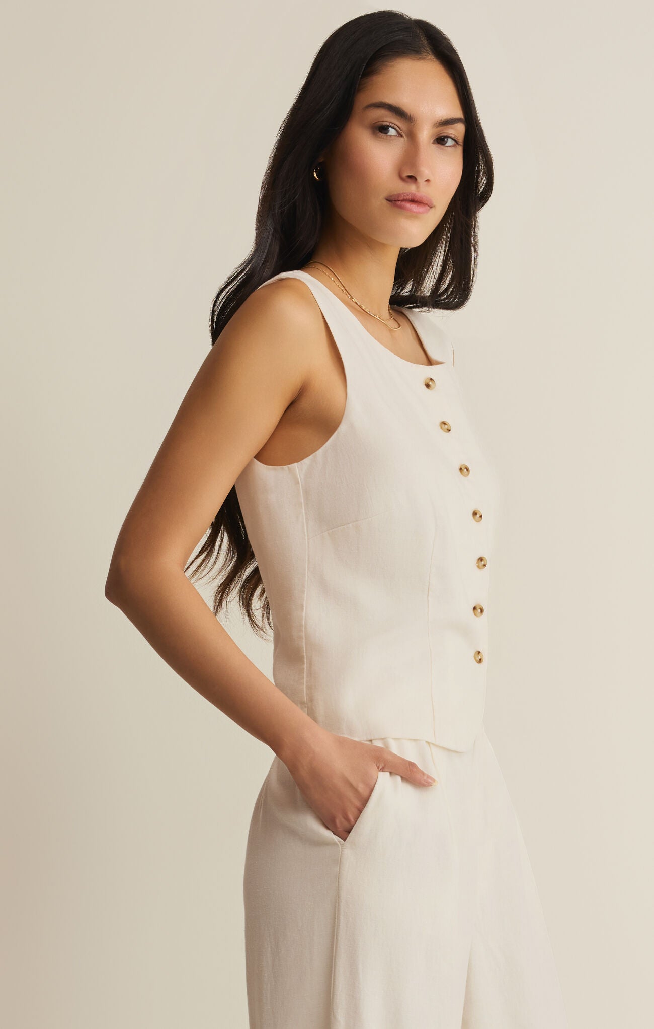 Woman wearing a white sleeveless top with gold buttons on a beige background