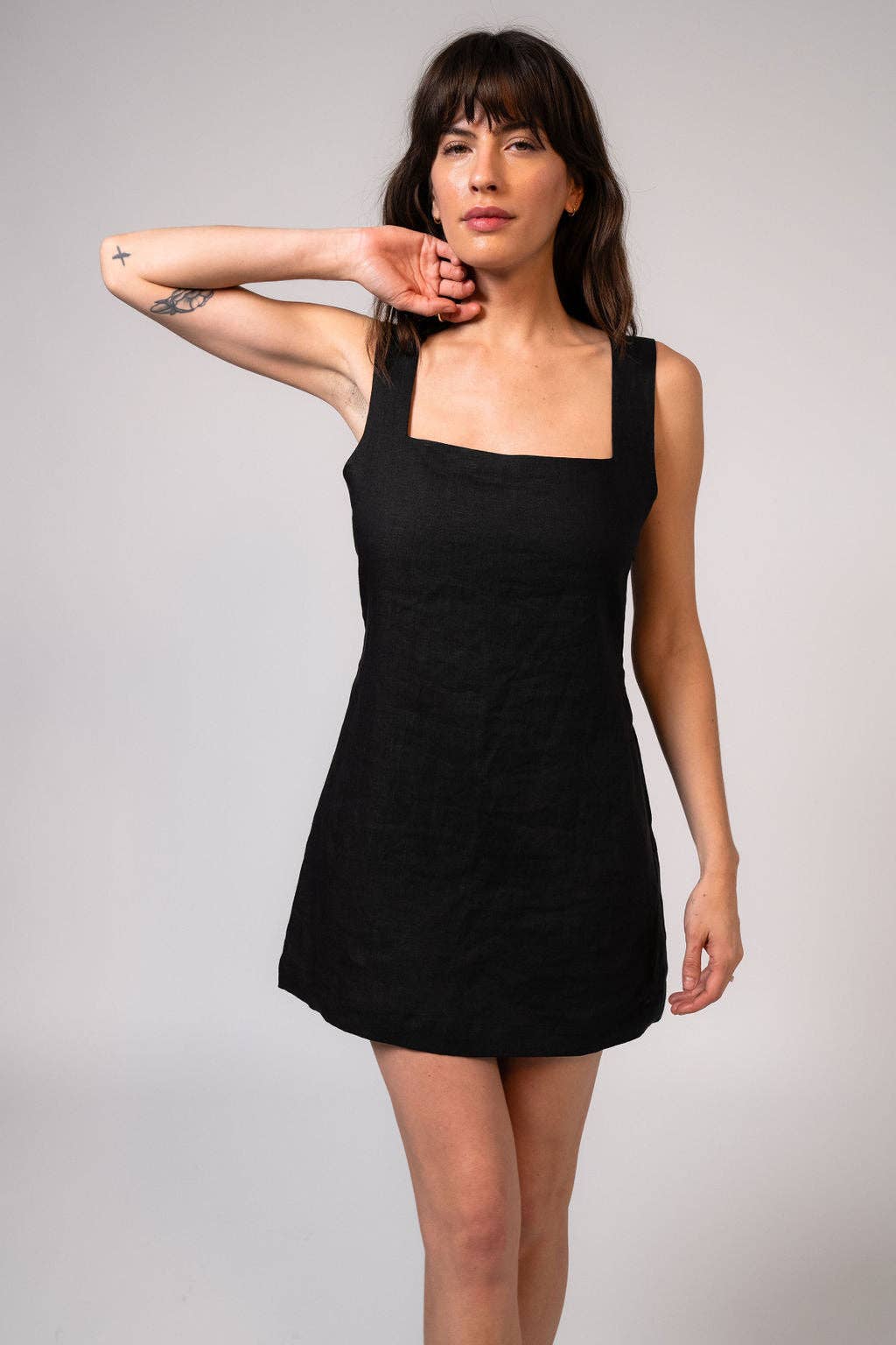 A woman modeling a black sleeveless mini dress with a square neckline and wide shoulder straps, posed against a plain light gray studio background.