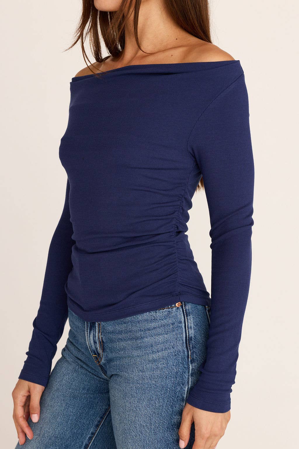A woman wearing a navy blue off-the-shoulder long-sleeve top with ruching on the side, paired with blue jeans, against a plain light beige background.