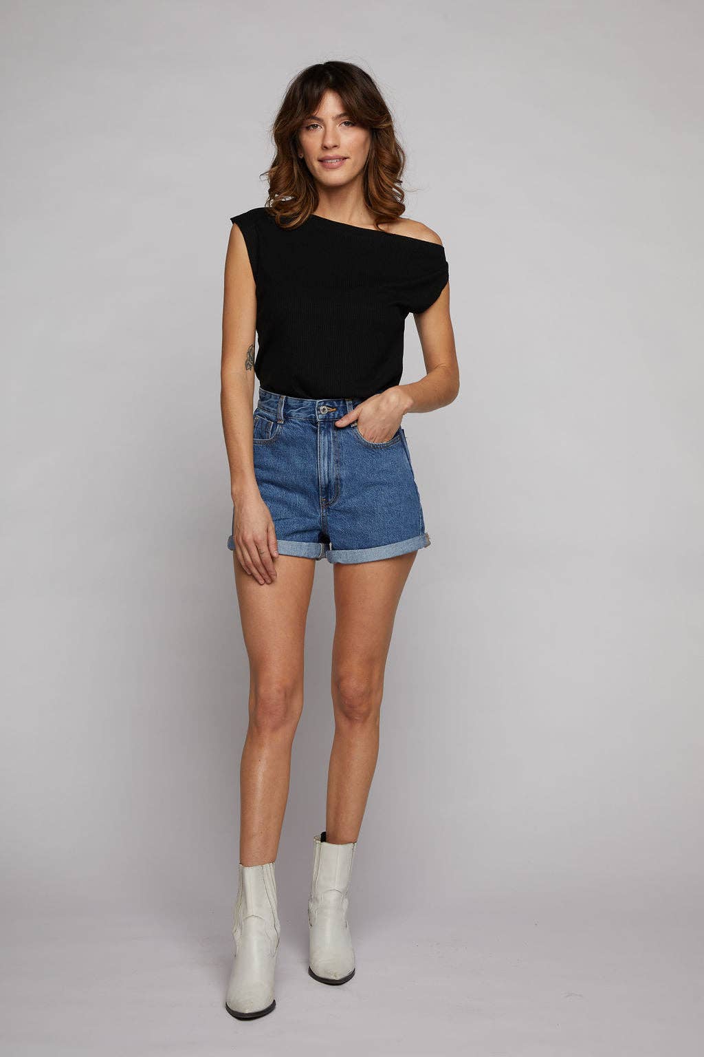 Woman wearing a black off-shoulder top, blue denim shorts, and white ankle boots on a gray background