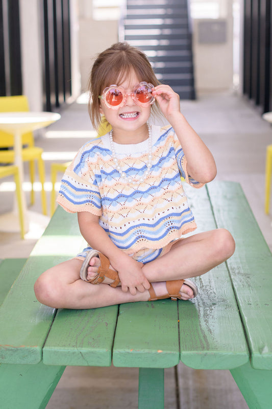 Young girl sitting on a green bench wearing sunglasses in an indoor setting.