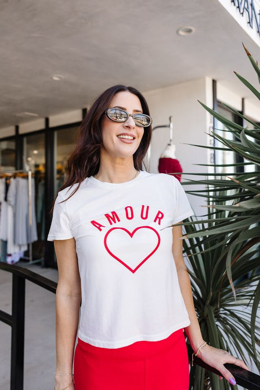 Woman wearing a white t-shirt with 'AMOUR' and a red heart design, standing outdoors.