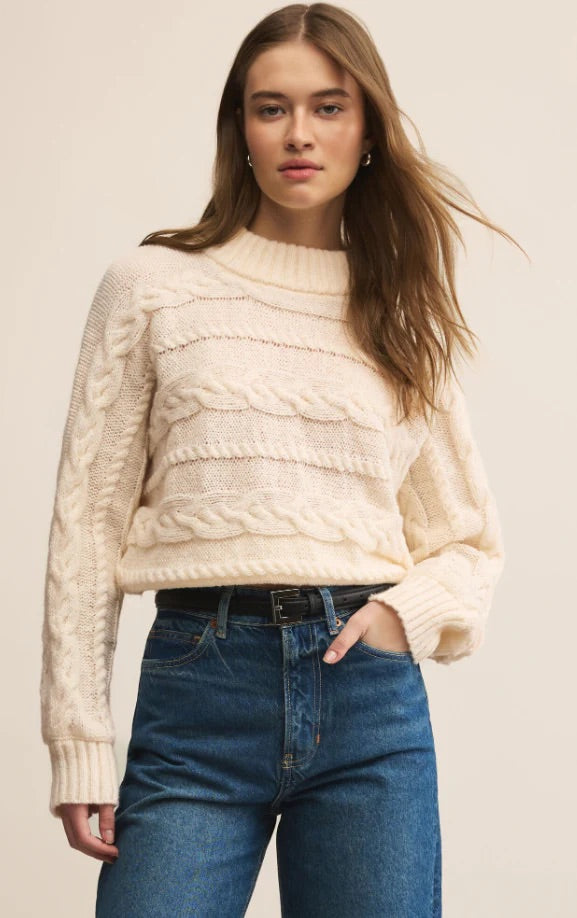 Woman wearing a cream cable knit sweater and blue jeans on a beige background