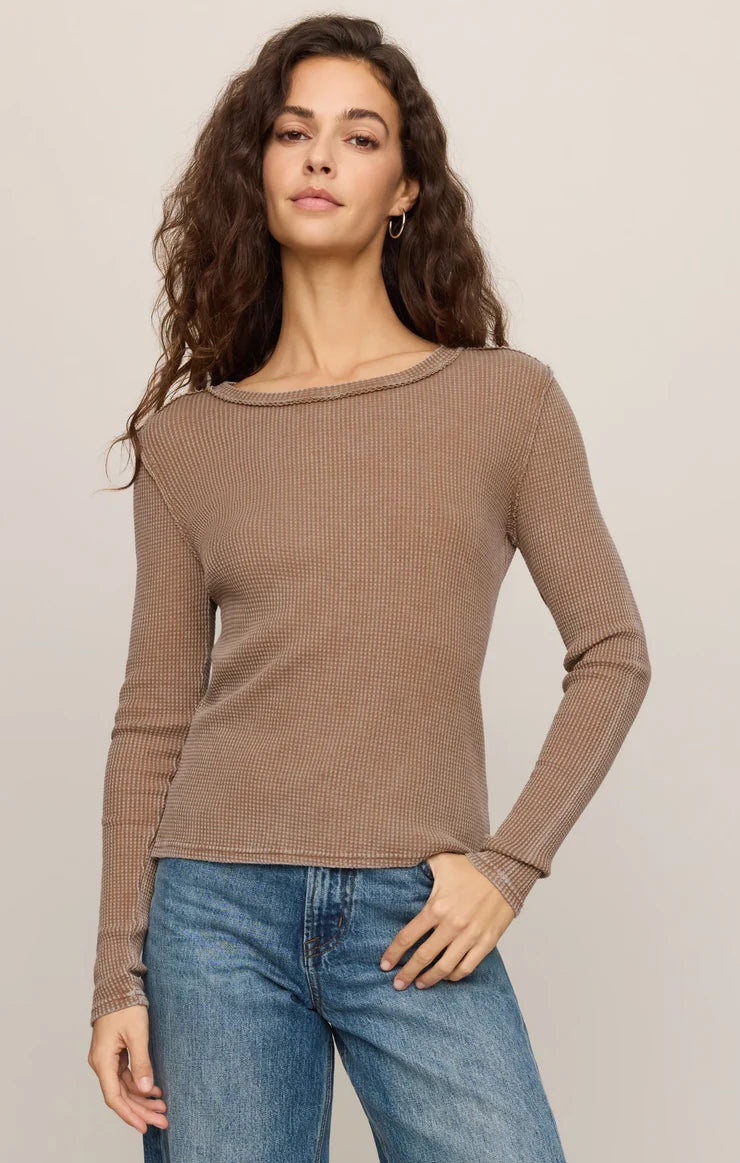 A woman modeling a fitted, long-sleeve ribbed knit top in taupe brown with a wide boat neckline, paired with blue jeans, against a solid light beige background.