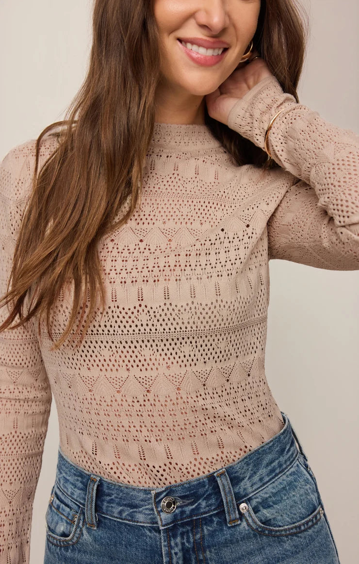 A woman wearing a beige open-knit long-sleeve mock-neck top paired with blue jeans, shown from the chest up against a plain light background.