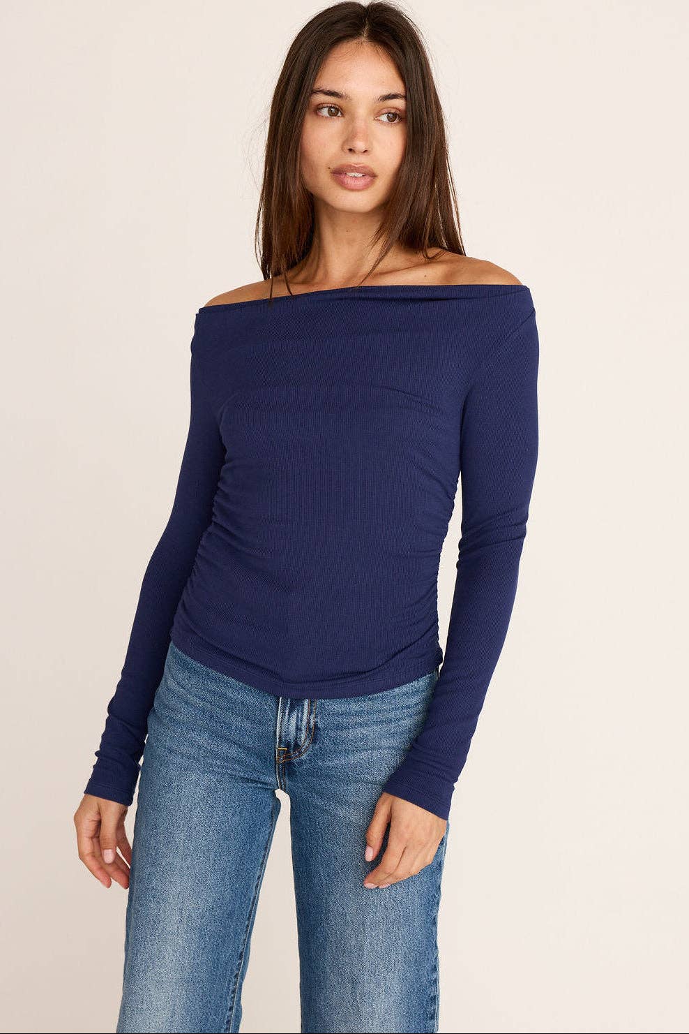 A woman modeling a navy blue off-the-shoulder long-sleeve top paired with blue jeans, photographed against a plain light beige studio background.