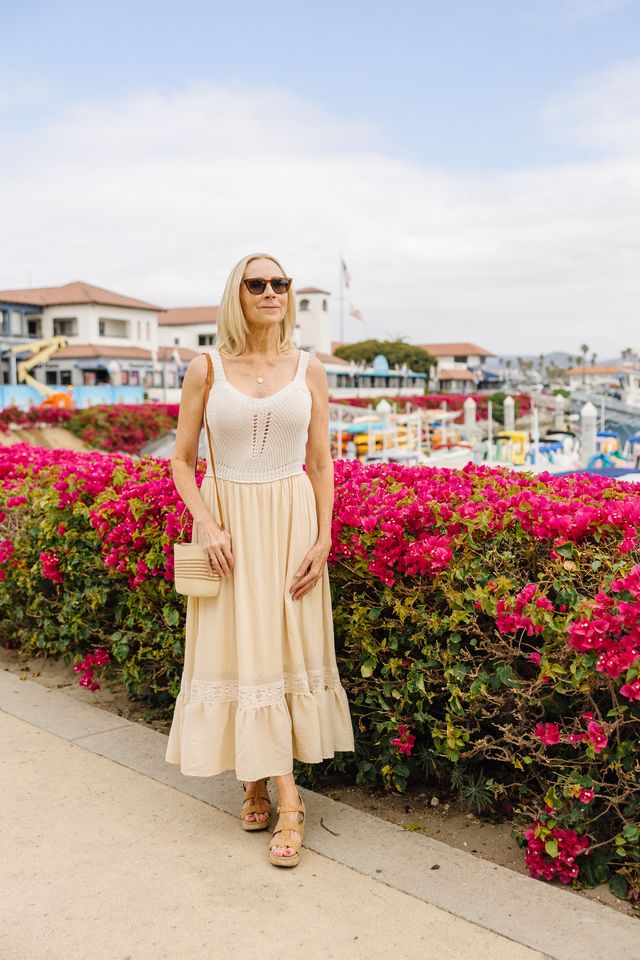 A woman wearing a cream crochet knit midi dress with a ruffled hem, standing outdoors on a boardwalk next to a vibrant pink bougainvillea hedge. She is holding a small beige woven clutch and wearing sunglasses and tan sandals.