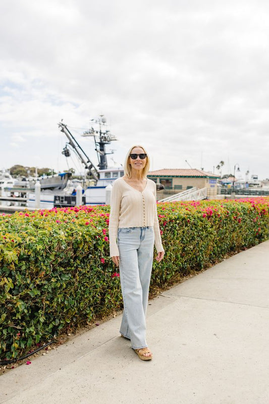 A woman wearing a beige knit cardigan, light-wash flared jeans, and sunglasses stands on a concrete walkway next to a green hedge with pink flowers, with boats and an overcast sky in the background.