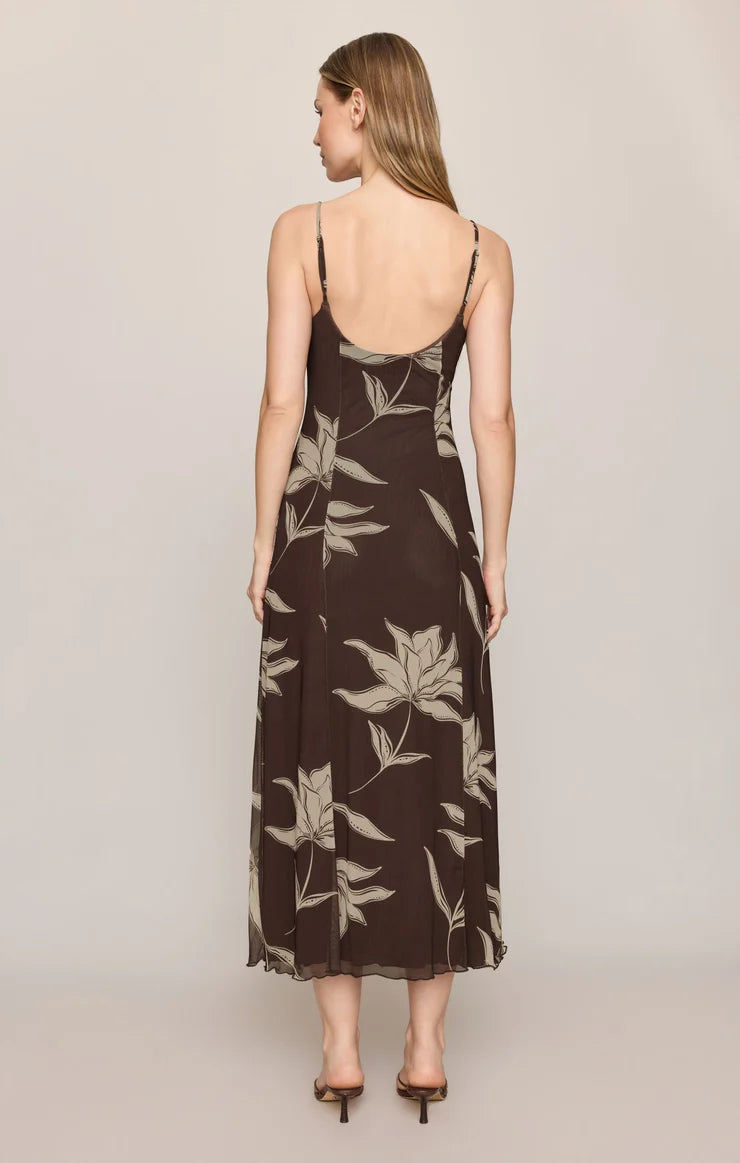 A woman viewed from the back wearing a sleeveless midi slip dress with thin straps and a brown tropical leaf print, paired with brown heeled sandals, against a solid light beige studio background.