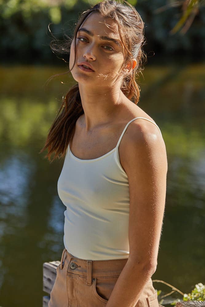 Woman wearing a white tank top and brown pants standing outdoors with a blurred natural background