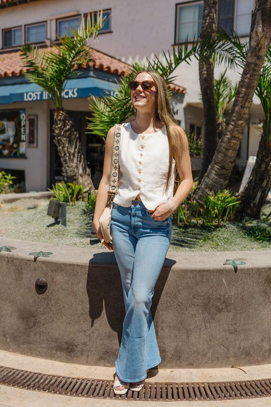 Woman in a white blouse and blue jeans standing outdoors with palm trees and a building in the background.