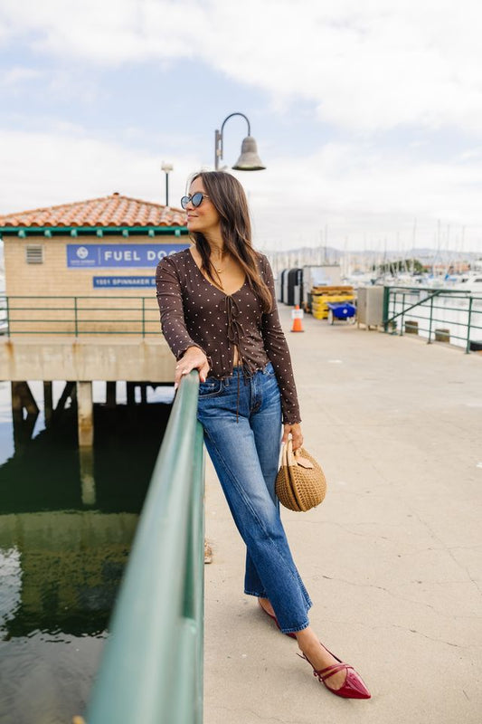 A woman stands on a sunny outdoor pier wearing a brown polka-dot long-sleeve crop top, blue jeans, burgundy pointed flats, and holding a woven straw bag.