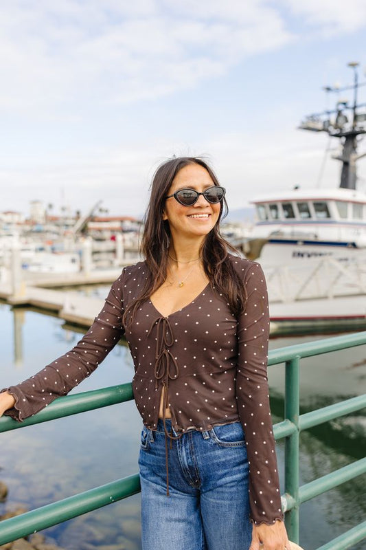A woman wearing a brown long-sleeve top with white polka dots and blue jeans stands by a green railing at a harbor, with boats and water in the background.