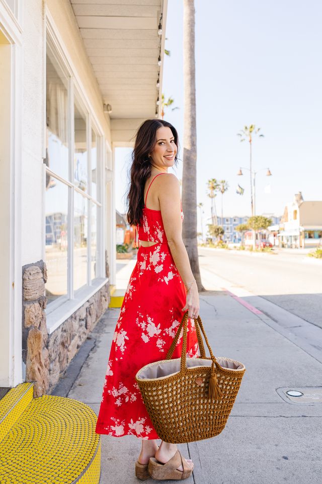 Woman in a red floral dress holding a straw bag on a sidewalk.