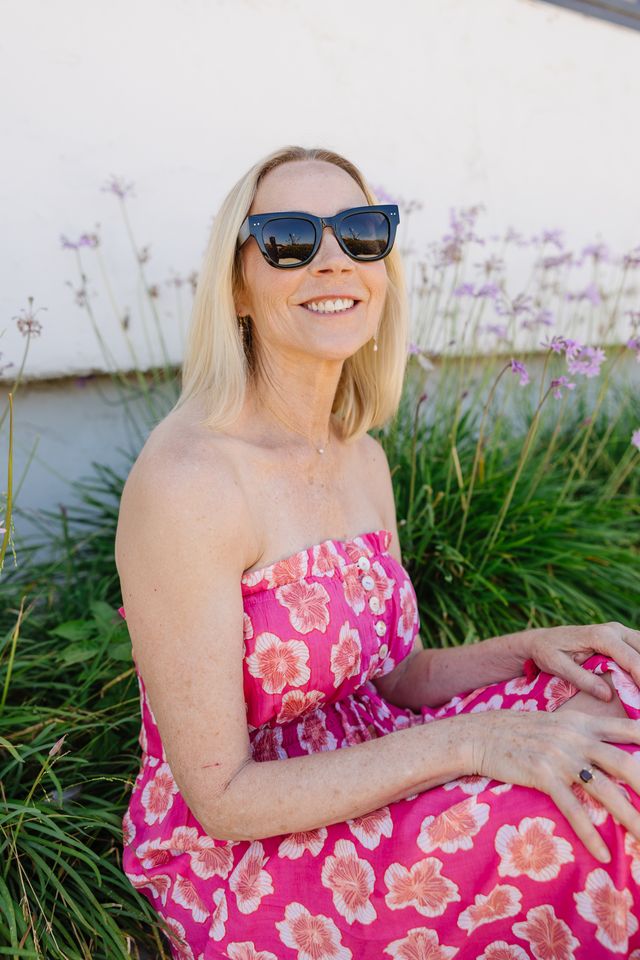 A smiling blonde woman wearing black sunglasses and a bright pink strapless dress with a large floral print, sitting outdoors among green plants and small purple flowers.