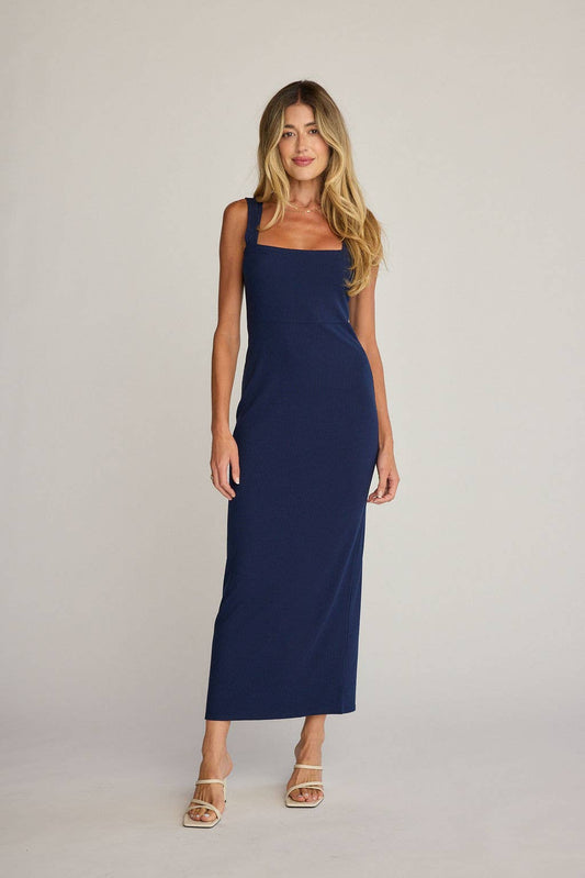 A woman modeling a sleeveless navy blue maxi dress with a square neckline, photographed against a plain light gray studio background.