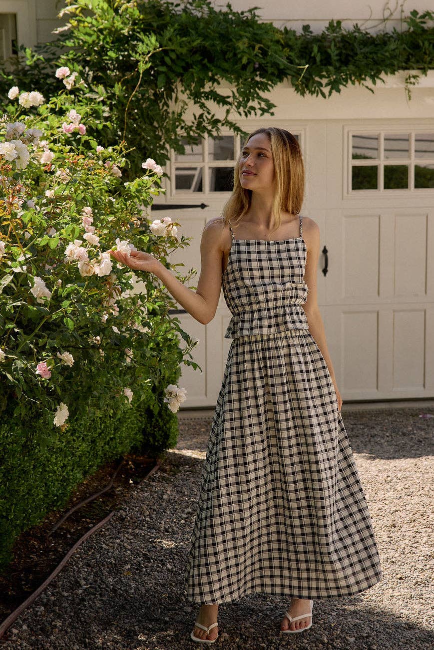 Woman in a checkered dress standing next to a flowering bush in front of a white building.
