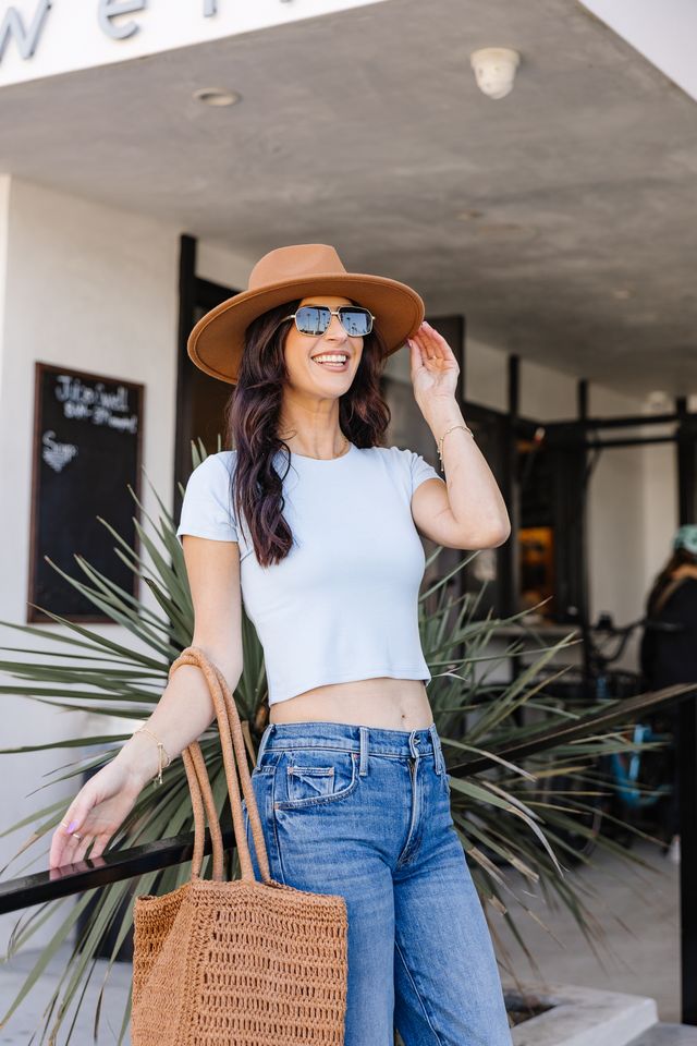 Woman wearing a hat and sunglasses, standing in front of a building with plants.