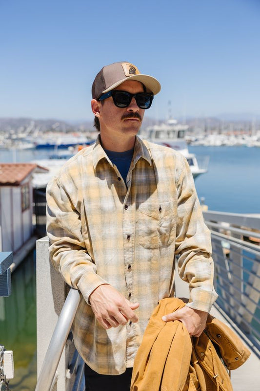 A man wearing a tan and beige plaid button-down shirt, sunglasses, and a baseball cap stands on a balcony overlooking a harbor with boats. He is holding a tan jacket.