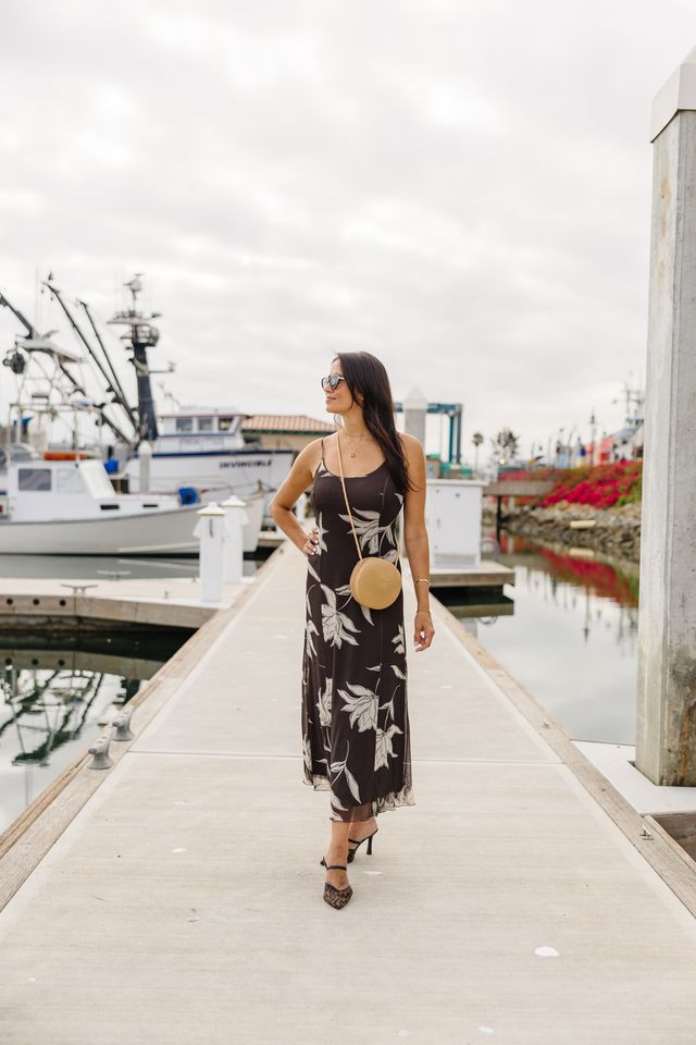 A woman wearing a black midi slip dress with a white tropical floral print walks along a concrete dock at a harbor. She is carrying a round beige straw bag and wearing black high heels.