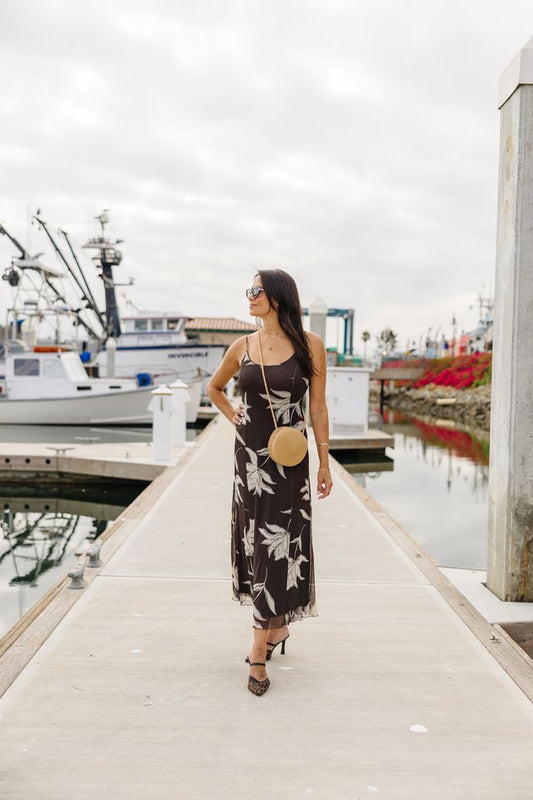 A woman wearing a black midi slip dress with a white tropical floral print walks along a concrete dock at a harbor. She is carrying a round beige straw bag and wearing black high heels.