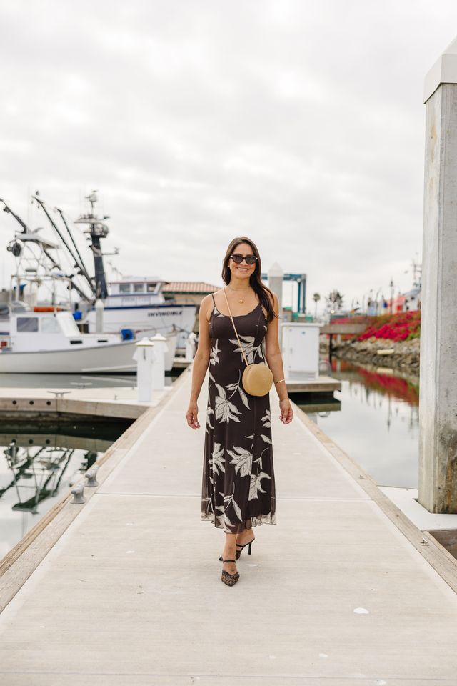 A woman wearing a dark brown sleeveless maxi dress with a white tropical floral print walks down a concrete pier at a harbor. She is carrying a round beige crossbody bag and wearing black high-heeled sandals.