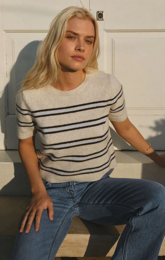 A blonde woman sitting outdoors against a white paneled wall, wearing a cream knit sweater with thin black horizontal stripes and blue jeans.