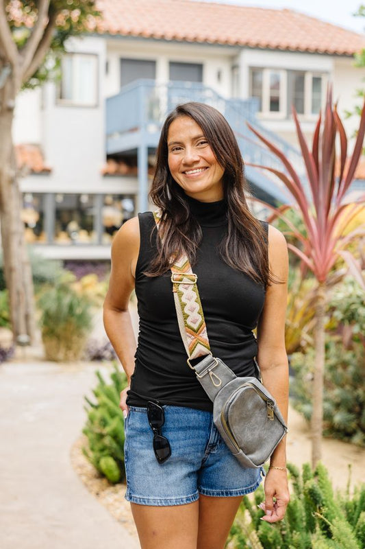 A woman wearing a black sleeveless mock neck top and denim shorts stands outdoors in a garden setting, wearing a gray crossbody bag with a patterned beige strap.