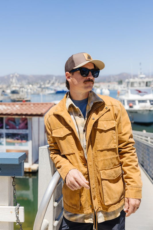 A man wearing a tan utility jacket, sunglasses, and a baseball cap stands on a dock with boats and water in the background.