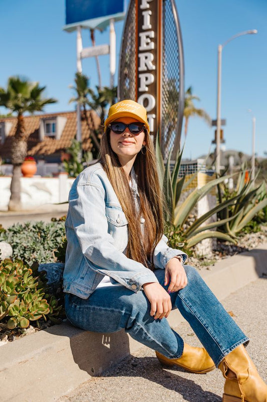Woman sitting on a bench wearing a denim jacket, yellow cap, and sunglasses with a pier sign in the background.
