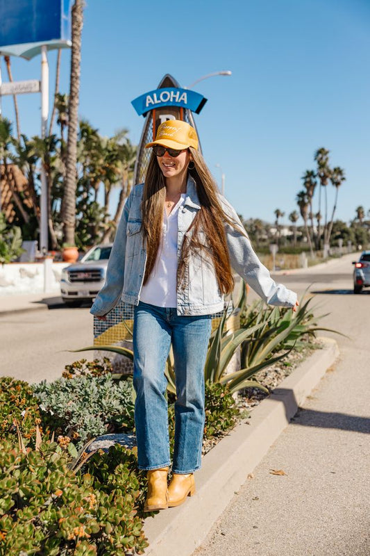 Woman wearing a yellow hat with 'ALOHA' text on a street with palm trees in the background