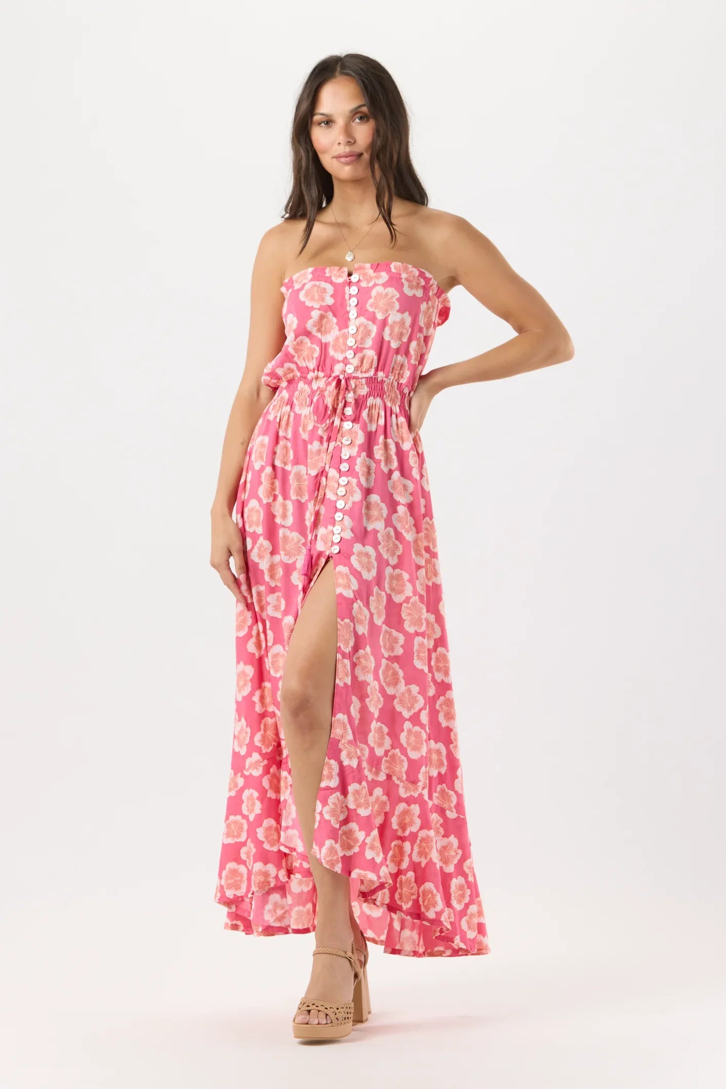 A woman modeling a strapless pink maxi dress with a white floral pattern and a high leg slit, paired with tan platform sandals, against a plain white studio background.