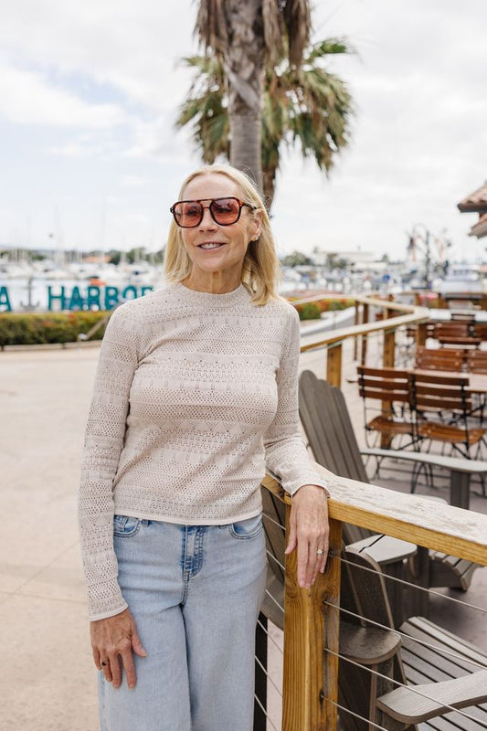 A woman wearing a beige textured knit mock neck sweater and light blue jeans stands outdoors on a boardwalk next to a wooden railing, with a marina and palm tree in the background.