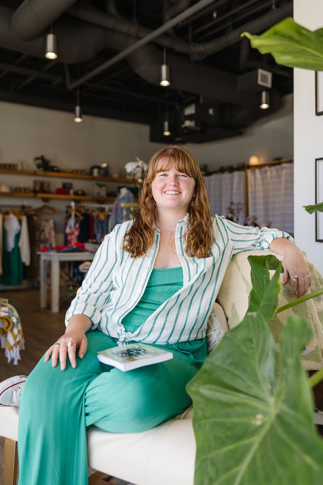 Woman sitting on a couch in a casual indoor setting with plants and shelves in the background.