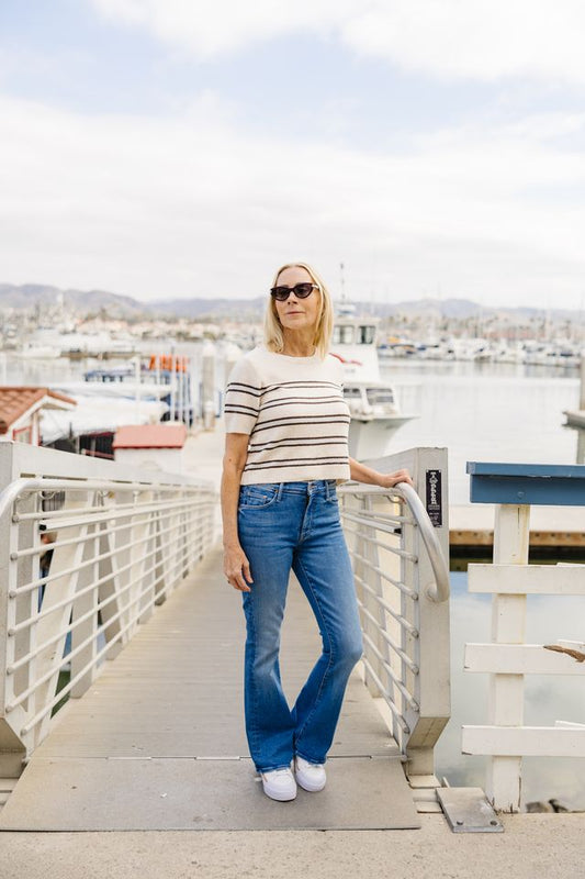 A woman stands on a wooden pier at a marina, wearing a beige and white horizontally striped short-sleeve top, blue flare jeans, and white sneakers.