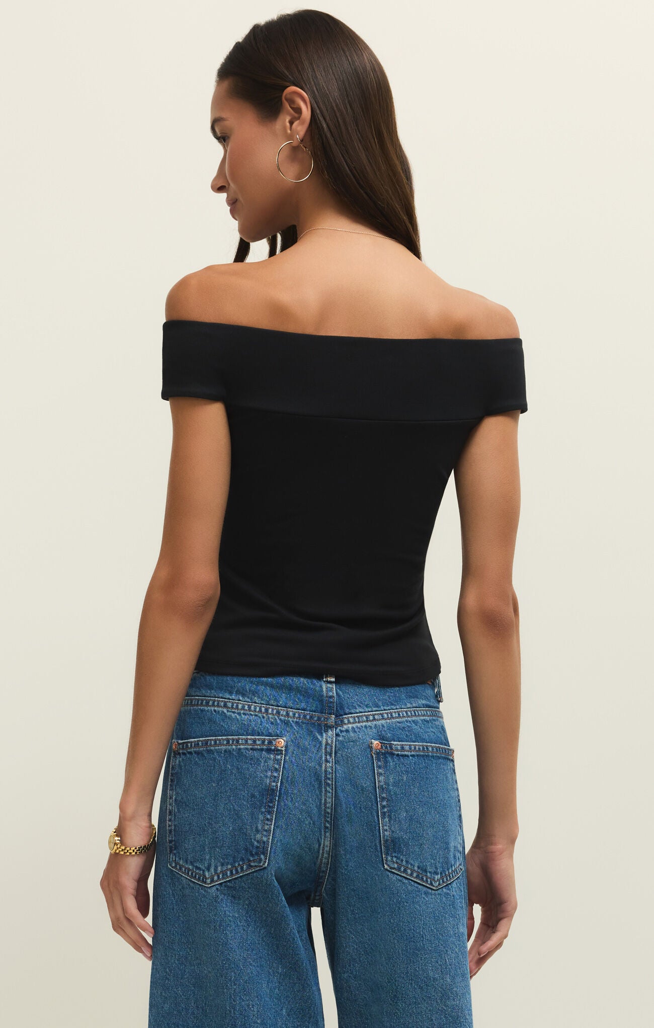 Woman wearing a black off-shoulder top and blue jeans on a beige background