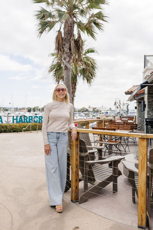 A woman stands on an outdoor waterfront patio wearing a beige ribbed long-sleeve top, light blue wide-leg jeans, and beige heels, with a palm tree and marina in the background.