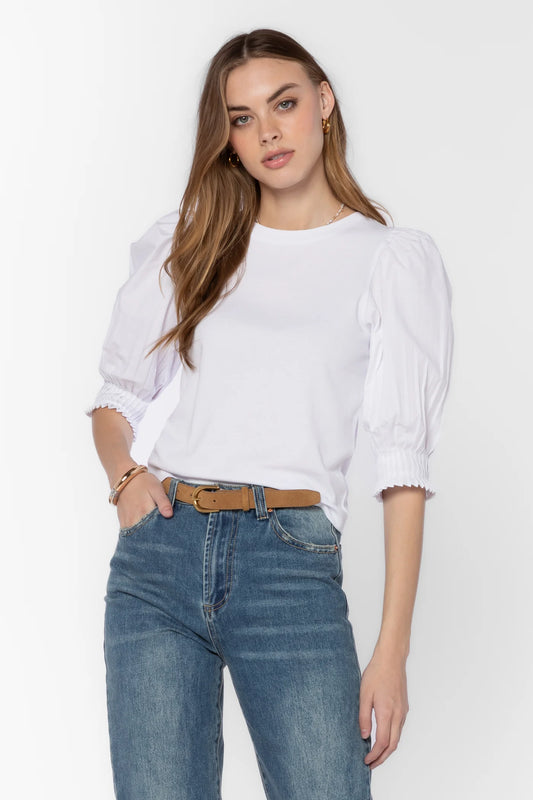 A woman modeling a white short-sleeve blouse with voluminous puffed sleeves and ruffled cuffs, paired with blue jeans and a brown belt, against a plain white studio background.