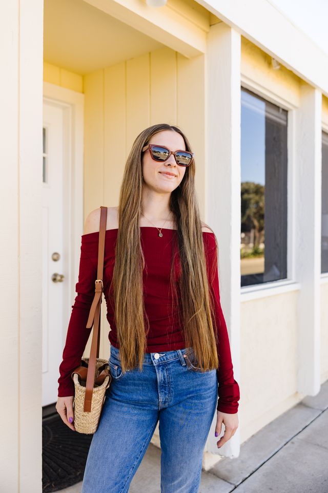 Woman wearing a red off-shoulder top and blue jeans standing in front of a house.