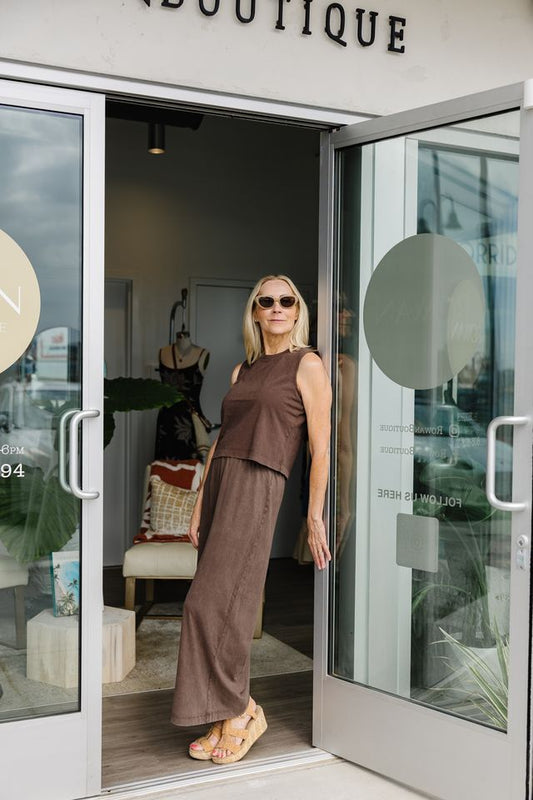 A woman wearing a brown sleeveless top and matching wide-leg pants stands in the doorway of a boutique, with the store interior and glass doors visible behind her.