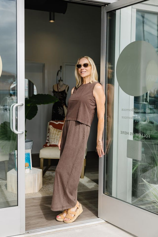 A woman wearing a matching sleeveless brown two-piece outfit with wide-leg pants and tan wedge sandals stands in a doorway of a boutique, smiling and posing.