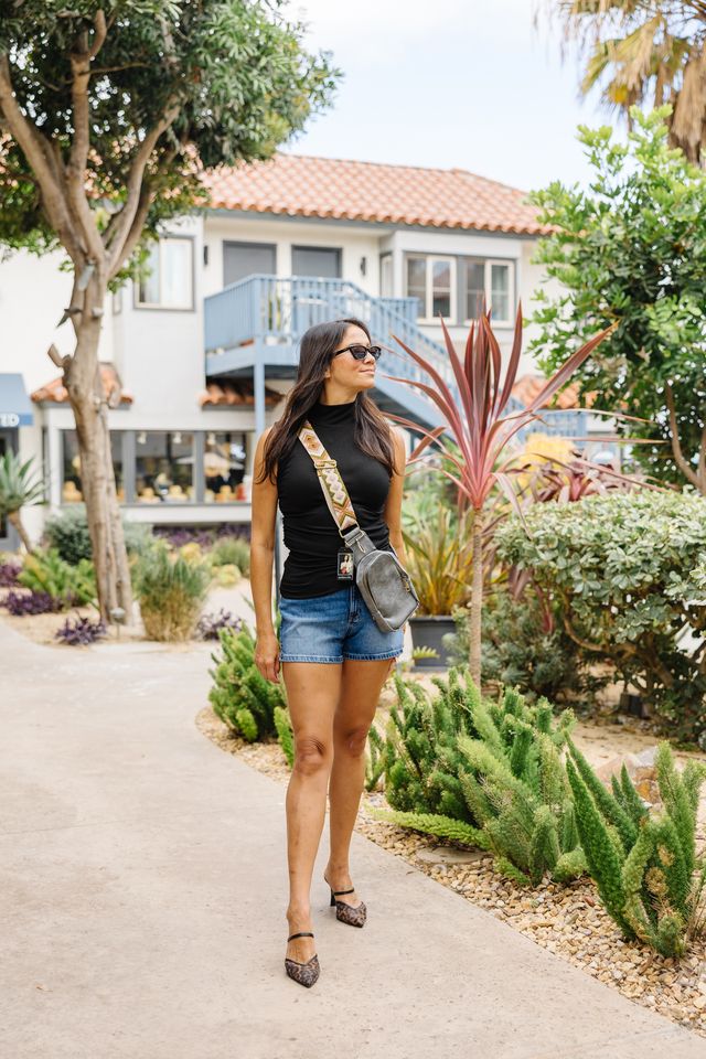 Woman in denim shorts and black high neck with greenery in the background.