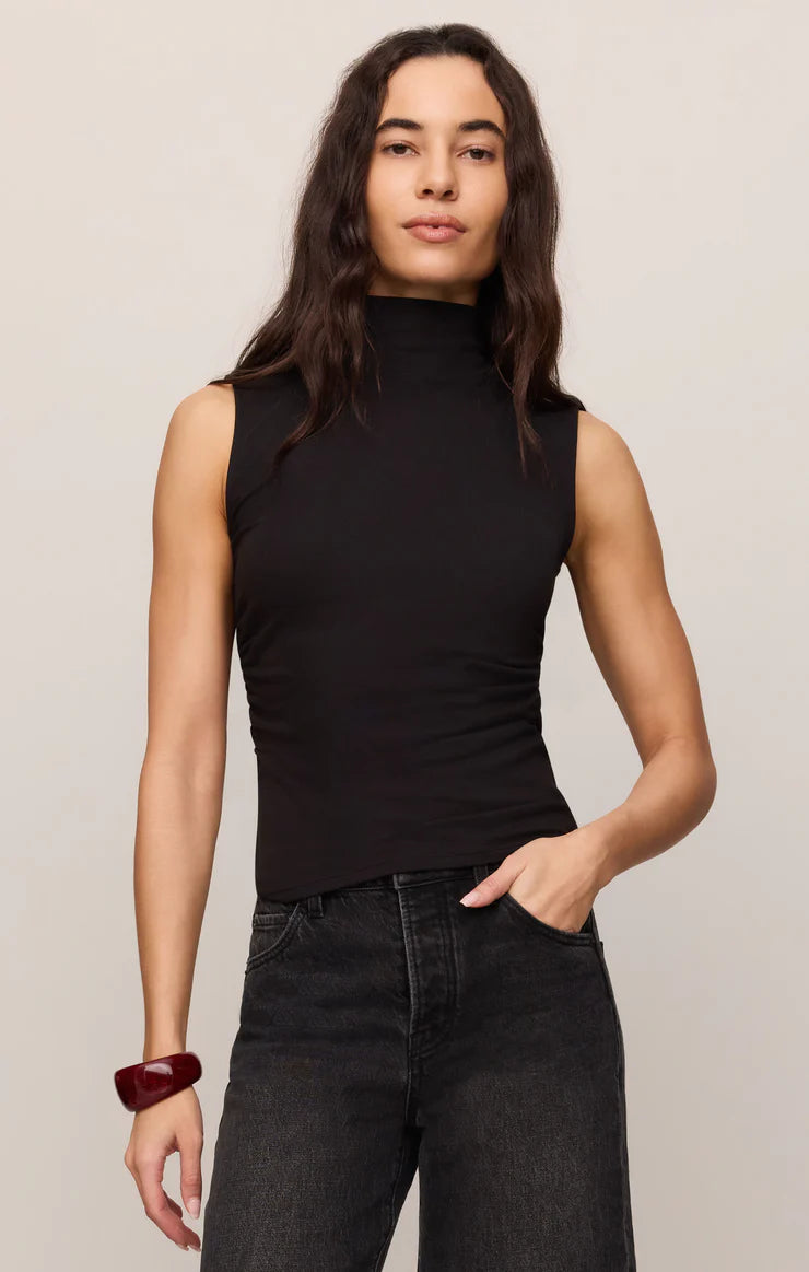 A woman modeling a sleeveless black mock neck top paired with dark gray jeans, standing against a plain light beige studio background.