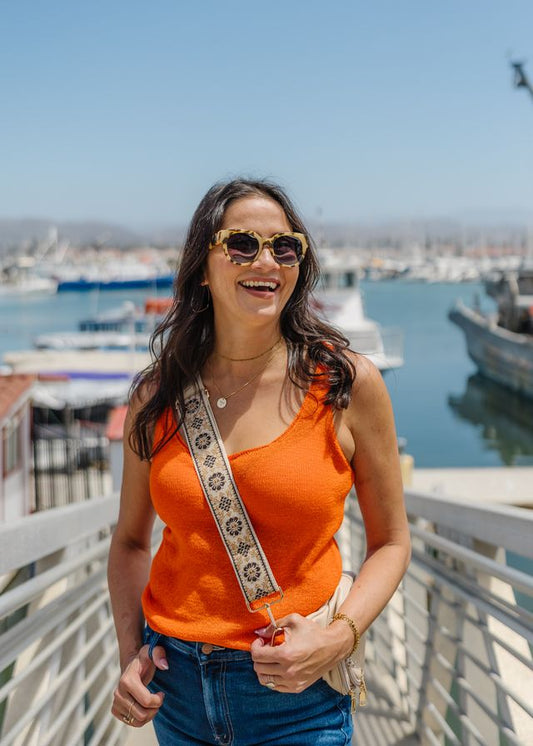 Woman in an orange top with a strap over her shoulder, standing by a waterfront.