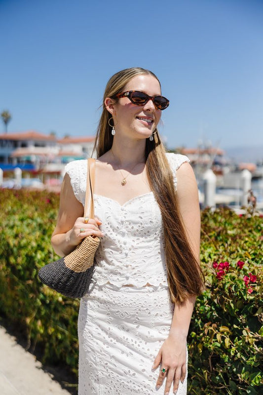 Woman in a white dress with sunglasses and a handbag standing outdoors with greenery and a clear blue sky.